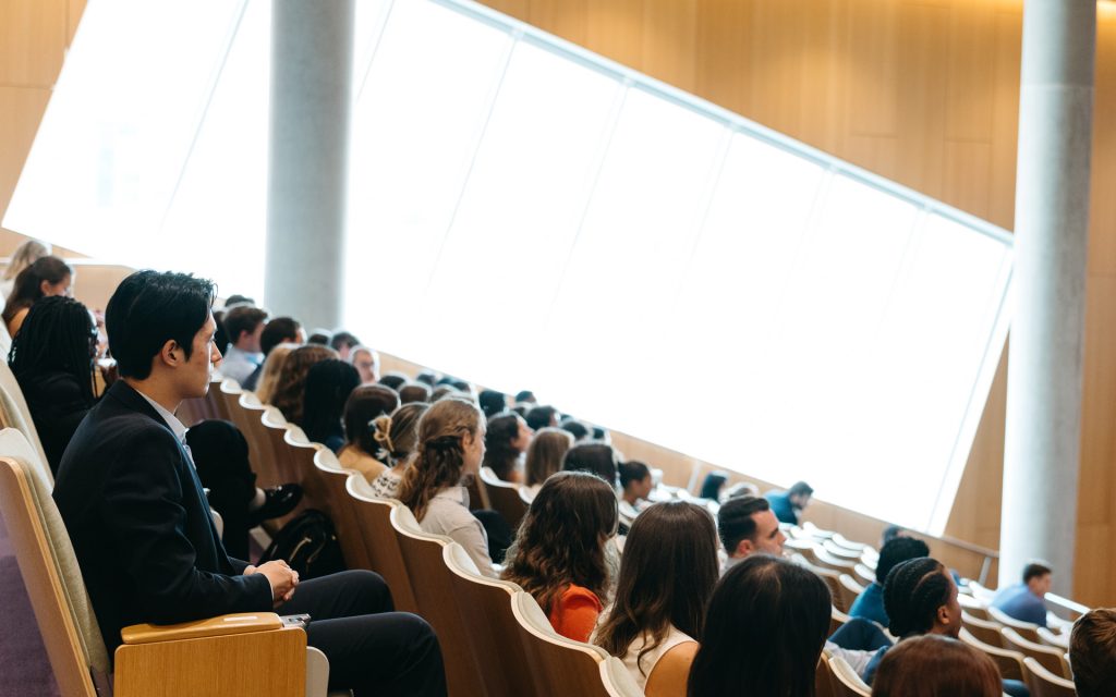 side view of a lecture hall audience, showing rows of participants attentively watching a presentation, with light streaming in from large windows.