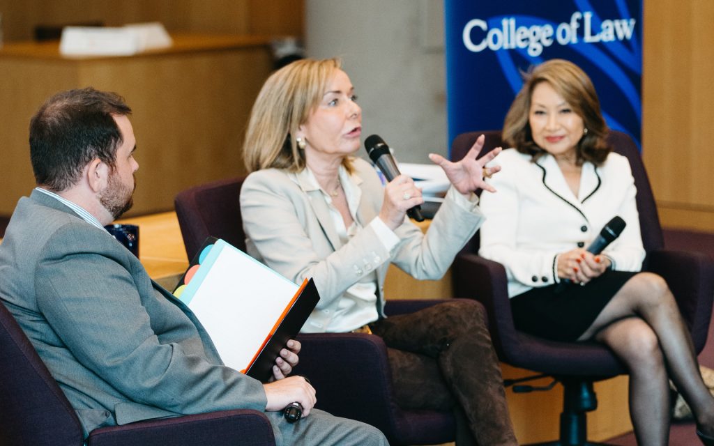 Three panelists engaged in a discussion on stage, with one woman gesturing expressively while speaking into a microphone. The backdrop includes "College of Law" branding.