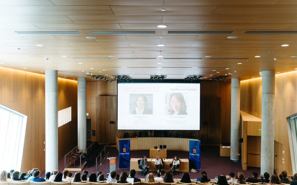 A wide-angle view of a lecture hall featuring a panel discussion on a stage, with a presentation slide displaying images of the panelists and their discussion topic, "Legal Competency in Tech."