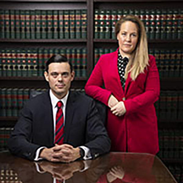 two lawyers, one male sitting at a desk with his hands crossed, and a female standing to his right in a red suit with her elbow on his shoulder, in front of law books