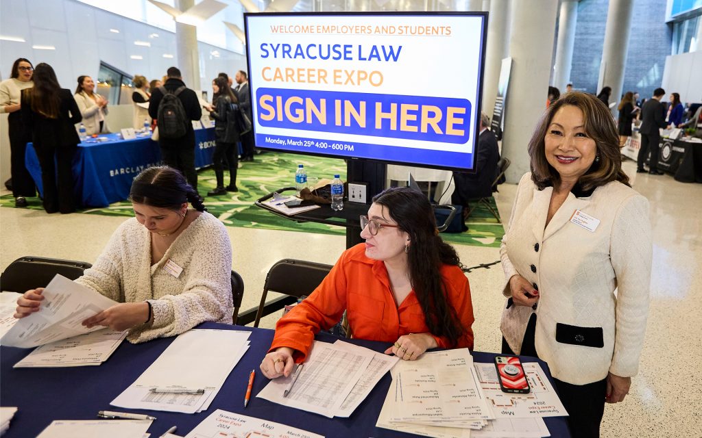 Two people working at a desk in front of a Syracuse Law Career Expo sign, a third person smiles at the camera