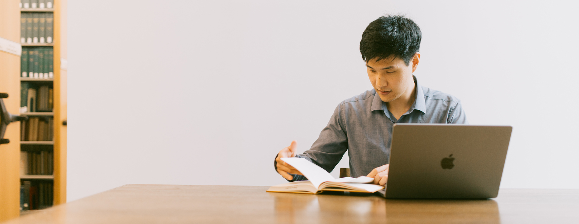 student reading a book with his laptop open in front of him on a desk in the library