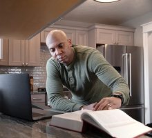 man looking at a law school book in his kitchen while also in front of his laptop