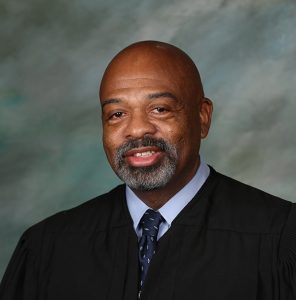 juge graves smiling at the camera in a black judge robe and a blue tie, in front of a gray abstract background