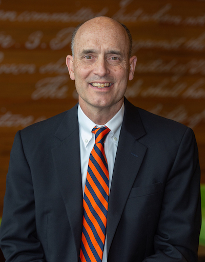 Judge James E. Baker, a bald, white man, wearing a black suit jacket over a white collared shirt with an orange and blue striped tie, smiles in front of an orange background.