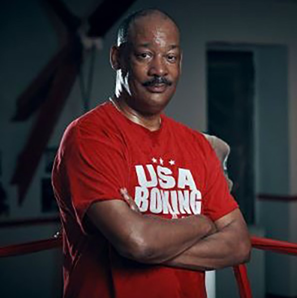 john elmore smiling in the corner of the boxing ring, with his arms crossed and a red usa boxing shirt on
