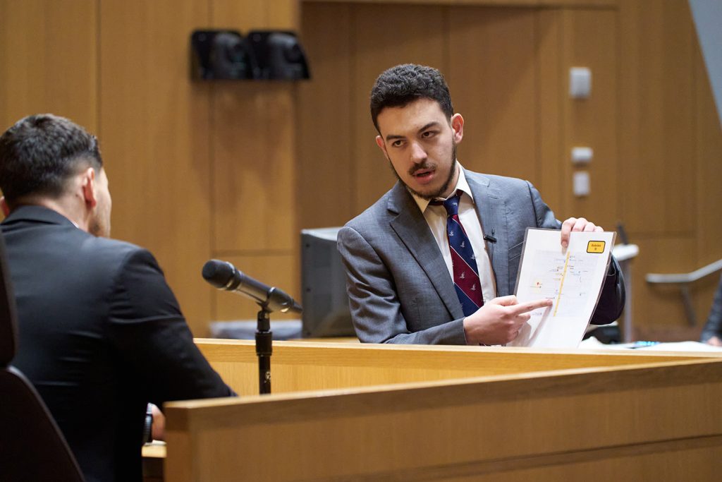 student points to a paper with evidence in a courtroom, standing in front of another person sitting with a microphone in front of him
