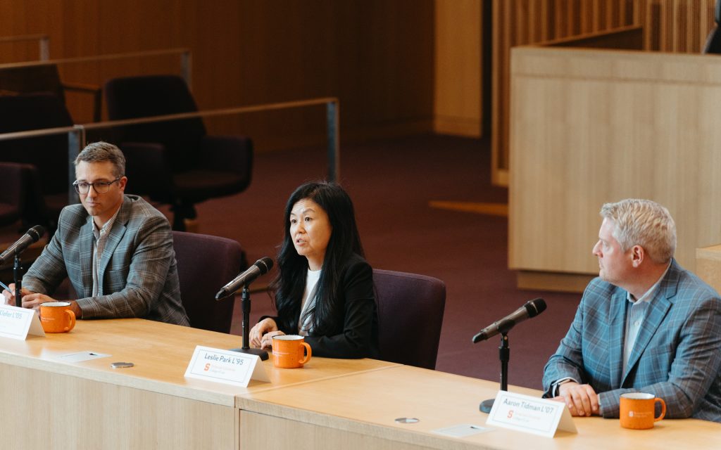 Three panelists seated at a table during a discussion, each with microphones and orange mugs. The middle speaker, a woman, is addressing the audience, while the two men on either side listen attentively.