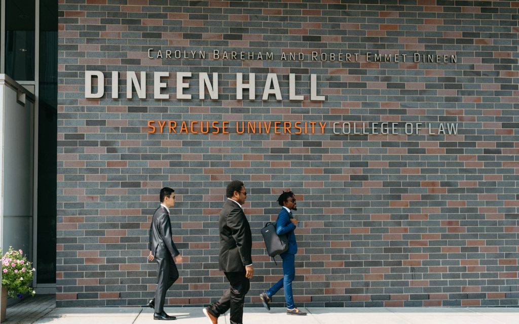 Three individuals in professional attire walking past the exterior of Dineen Hall, Syracuse University College of Law, with the building's name prominently displayed on the brick wall.