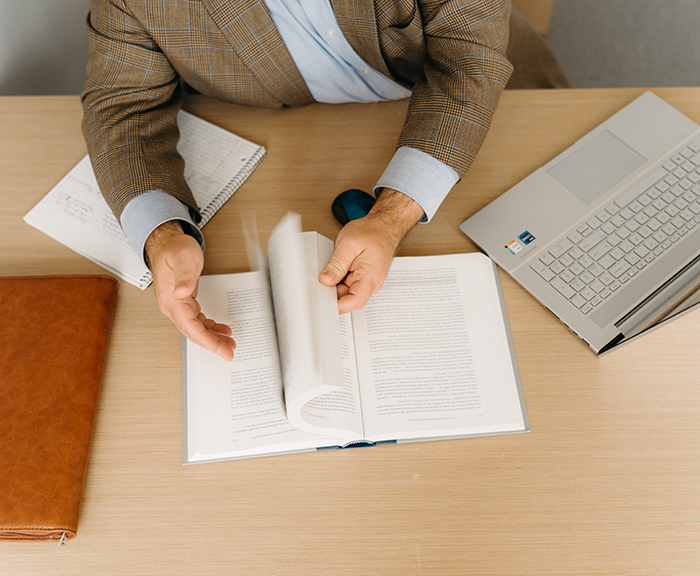 Abstract photo of man's hands leafing through a book and a laptop and leather briefcase on a desk in front of him.