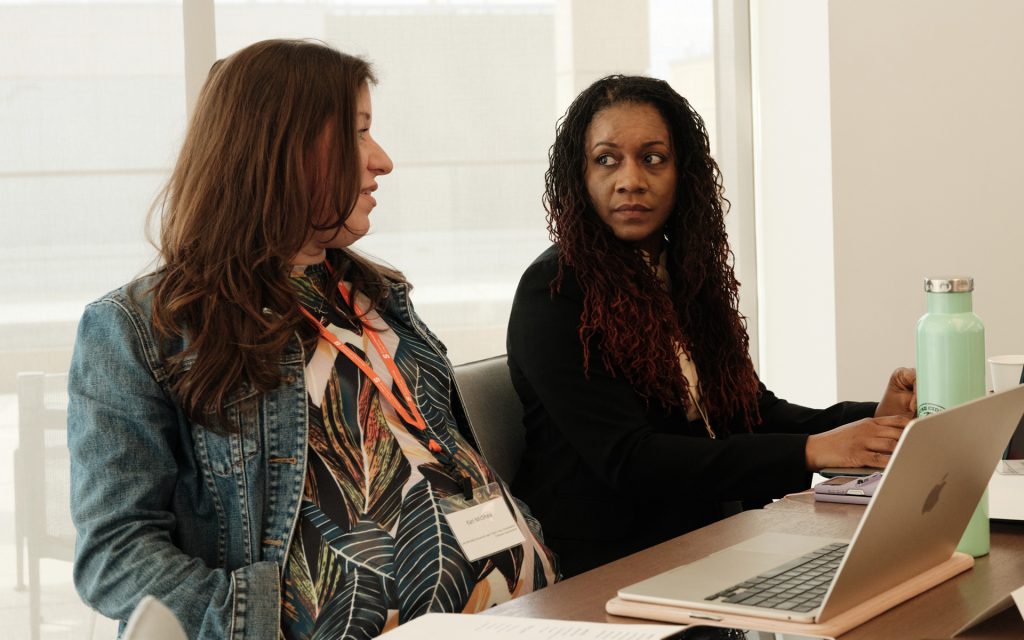 Two women sit at a table during a discussion or meeting. One wears a denim jacket over a patterned blouse, while the other, with braided hair, is dressed in formal attire. A laptop and a water bottle are visible on the table. Sunlight streams in through large windows in the background.