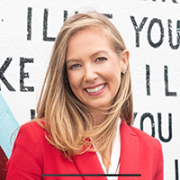 megan thomas smiling at the camera in front of a graffiti wall with the words I like You, wearing a red blazer