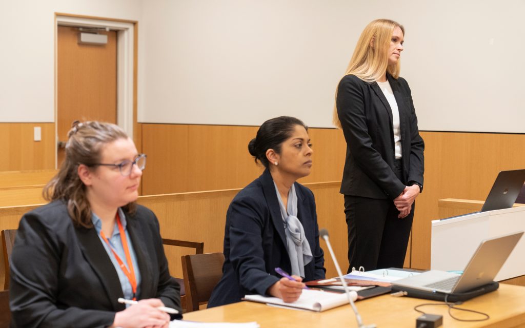 Three women participating in a courtroom simulation. Two women, seated at a table, wear professional business attire, focusing on their notes. One stands next to them, presenting with a laptop nearby. The courtroom setting has light wood paneling and neutral walls.