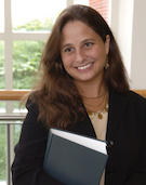 Professor Nina Kohn, a white woman with brown shoulder-length hair, wearing a black blazer over a tan sweater, with gold necklaces and earrings, smiles in front of a window. She is holding a dark blue book.