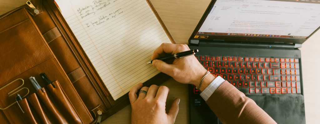 close-up of a leather portfolio, male hands writing on paper, and an open laptop on a desk