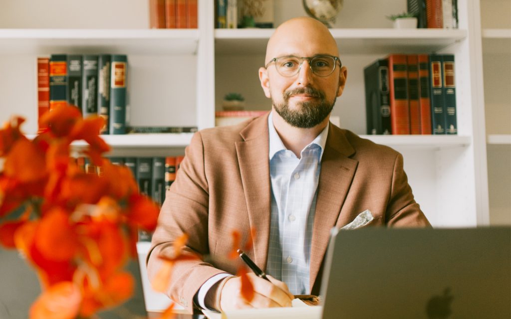 A man seated at a desk, writing in a notebook with a laptop in front of him. He is wearing glasses, a light blue shirt, and a brown blazer, with shelves of books and a globe visible in the background. Bright orange flowers are in the foreground, slightly blurred.