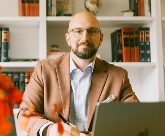 A man seated at a desk, writing in a notebook with a laptop in front of him. He is wearing glasses, a light blue shirt, and a brown blazer, with shelves of books and a globe visible in the background. Bright orange flowers are in the foreground, slightly blurred.