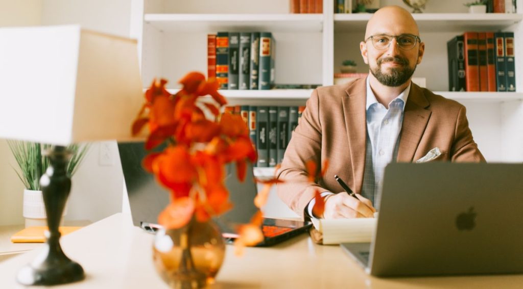 A man with a beard and glasses in a light orange suite in front of a computer with a pen in his hand.