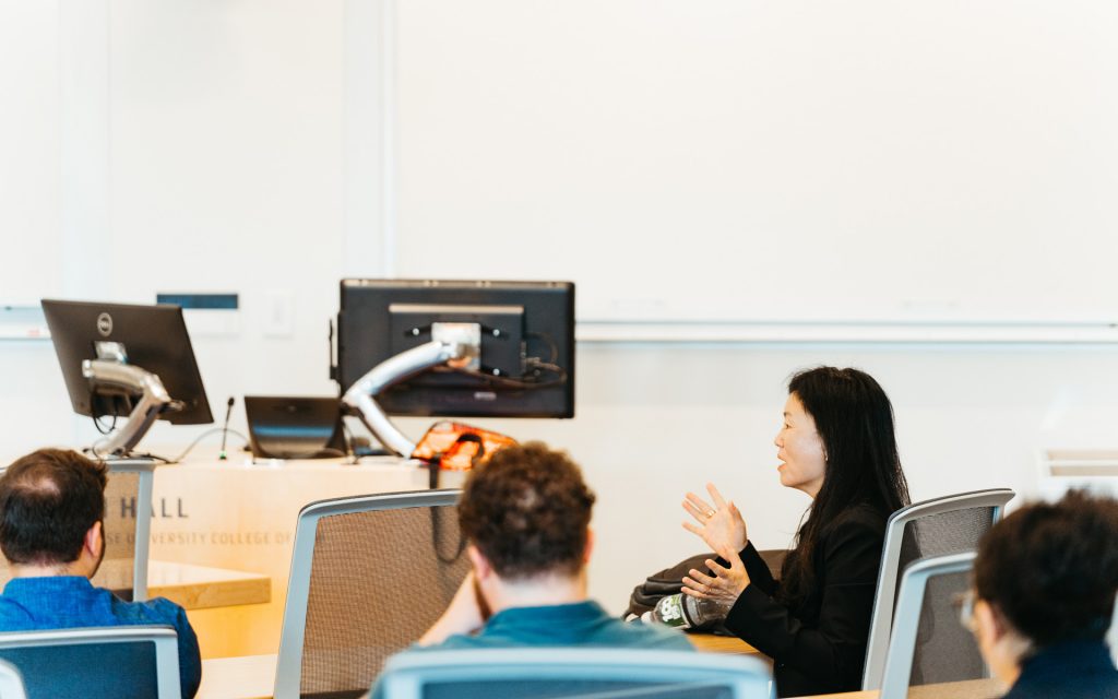 woman speaking and gesturing during a classroom session. The back of the audience’s heads and a computer setup on a podium are visible in the foreground.