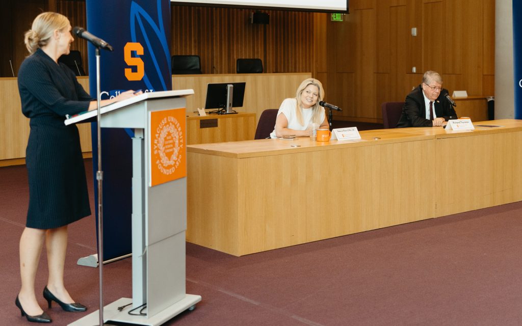 A woman standing at a podium presenting in a lecture hall with "Syracuse University" and "College of Law" branding. Two panelists seated at a table listen and smile during the presentation.