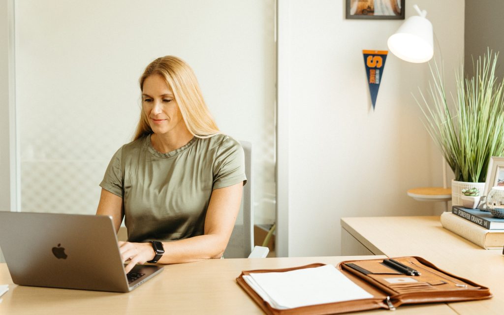 A woman in a green blouse, seated at a modern desk, working on a laptop in a bright office. The desk has a notebook, pen, and a Syracuse University pennant in the background, along with a decorative plant and books.