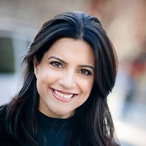 Reshma Saujani wearing a black shirt and smiling at the camera in front of a blurred-out background