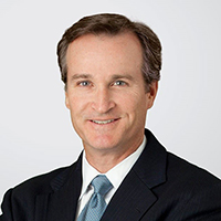 richard furey smiling at the camera in front of a white background and wearing a black suit and blue tie