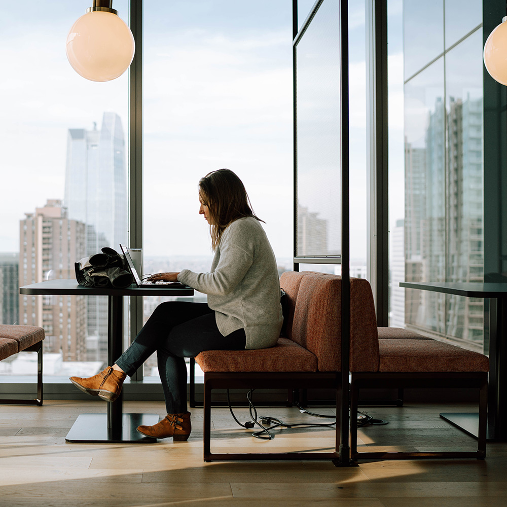 woman sits at a desk and works in front of a window with the buildings of Denver in the background