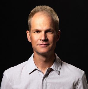 headshot of rob beard wearing a gray button down in front of a solid black background