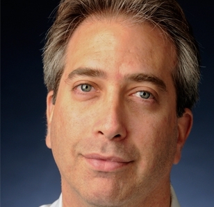 Professor Roy Gutterman, a white man with brown and gray hair, wearing a white collared shirt, smiles in front of a black background.