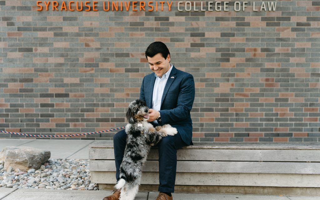 The man is sitting on a bench outside Syracuse University College of Law's Dineen Hall, smiling as he interacts with a small, fluffy dog standing on its hind legs with its paws resting on his lap.