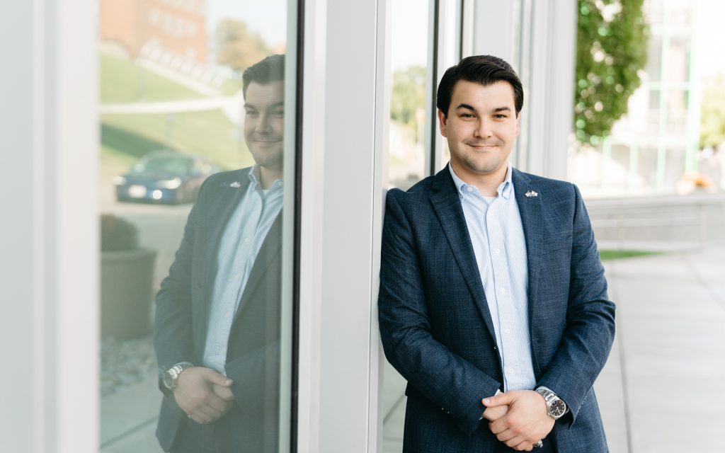 The man stands leaning against a glass wall at Syracuse University College of Law. His reflection is visible in the glass, and he is smiling slightly, dressed in a blue blazer and white shirt.