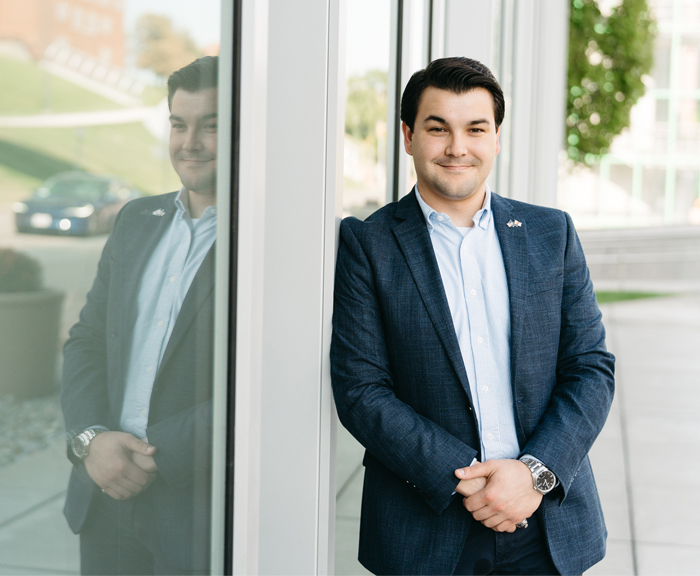 The man stands leaning against a glass wall at Syracuse University College of Law. His reflection is visible in the glass, and he is smiling slightly, dressed in a blue blazer and white shirt.