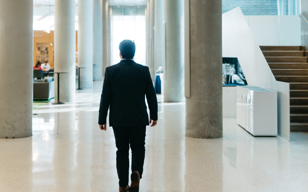 A view of the man from behind as he walks down a hallway inside Dineen Hall. The modern, open space is well-lit, with tall concrete columns and a polished floor.