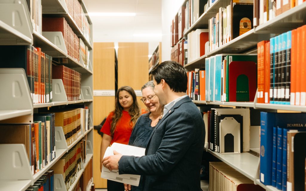 The man is standing between library shelves in Dineen Hall, looking at an open book with two other people nearby, engaging in a conversation and smiling.