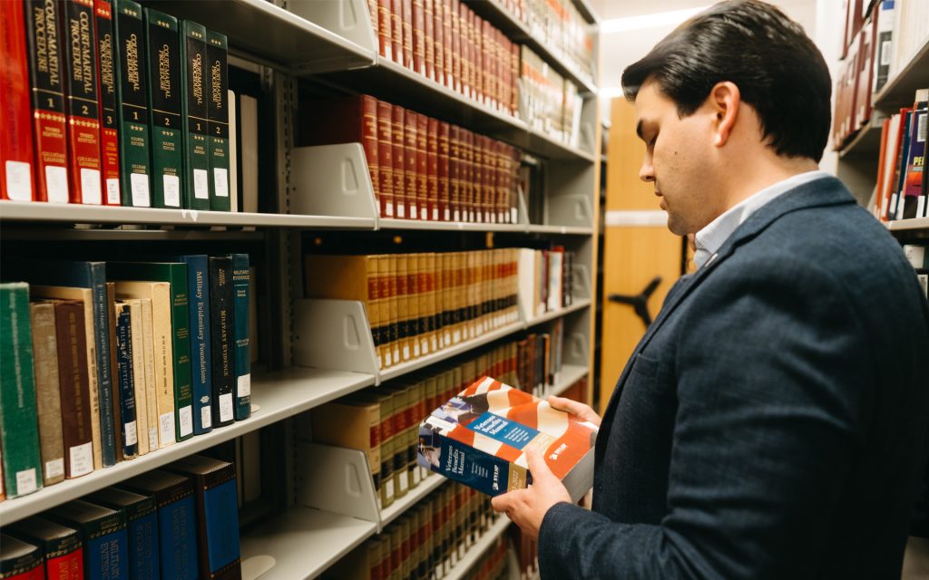 The man stands in a library aisle, examining a large law book. Rows of books on legal topics are visible on the shelves around him. He is dressed in a blue blazer and focused on the book in his hands.