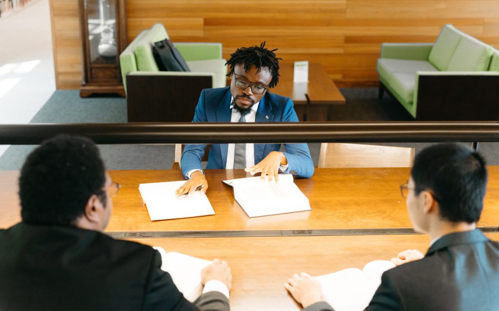 A group discussion in a library setting, with the student in a blue suit leading the conversation, gesturing to a book while two other individuals listen attentively.