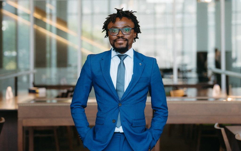 A portrait of the student in a blue suit, standing confidently with hands in pockets, in a well-lit modern library space.