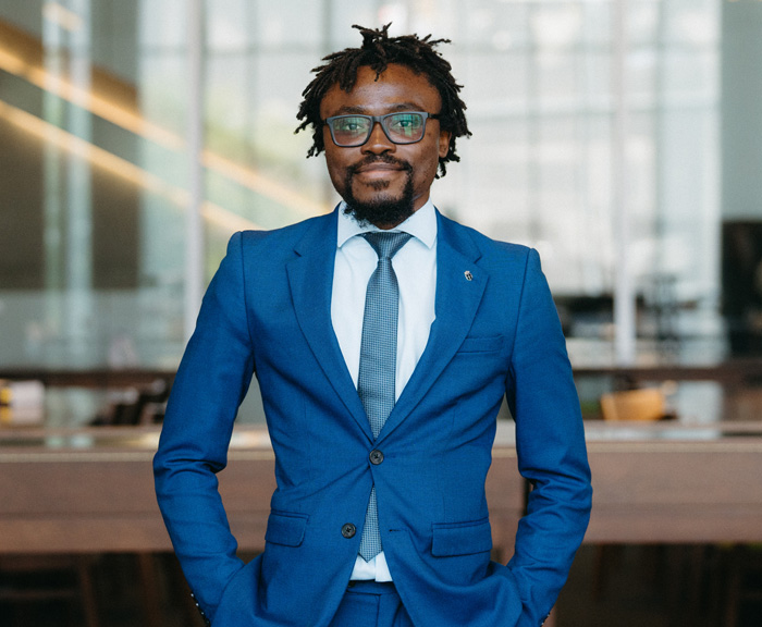 A portrait of the student in a blue suit, standing confidently with hands in pockets, in a well-lit modern library space.