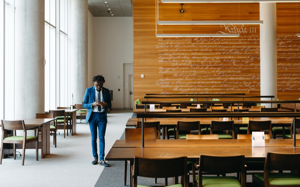 The student in a blue suit walking through the library, looking down at a phone, with the text of Article III of the U.S. Constitution displayed on the wood-paneled wall behind.