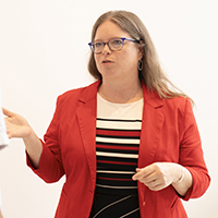 sarah reckess talking to a student, wearing a red blazer and black rimmed glasses in front of a white background