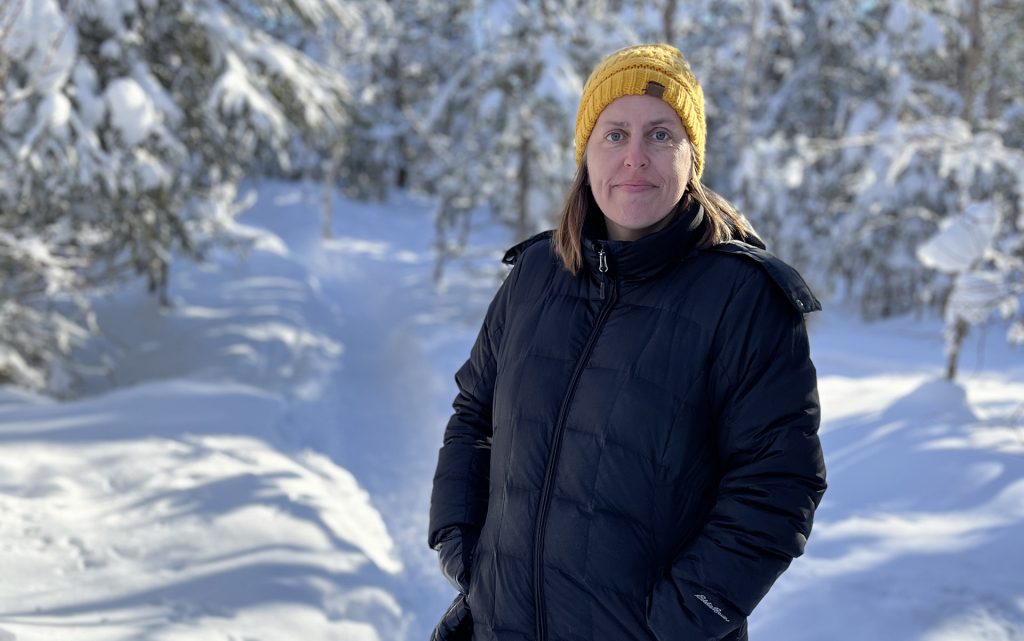 Sarah Roberts stands in a snowy field with evergreen trees in alaska, wearing a black coat and a yellow yat.