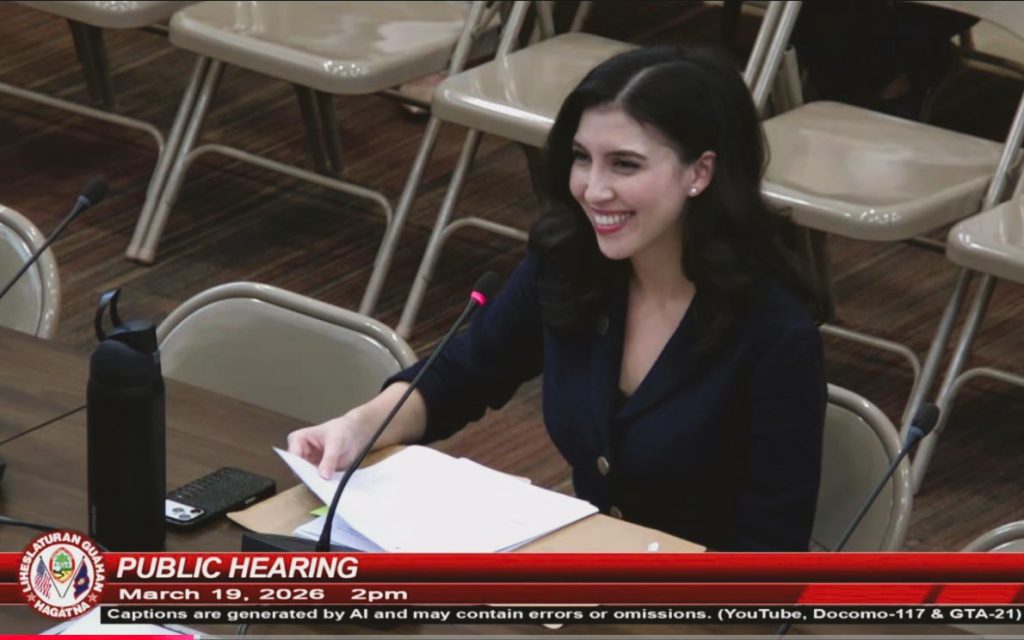 Elmore-Hernandez's shown on TV smiling and speaking into a microphone at a public hearing.