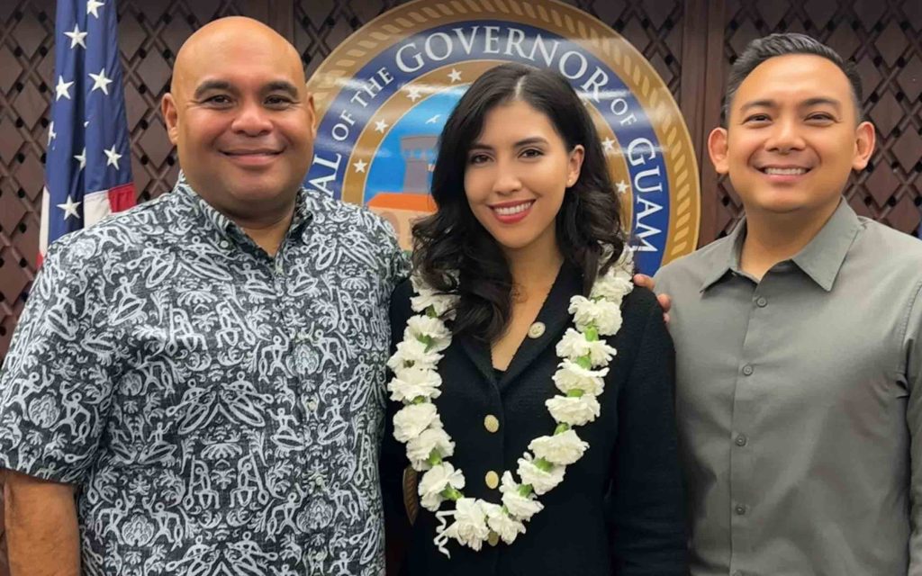 Sarah wears a lei necklance and smiles standing between two men in front of the Guam seal and US flag.