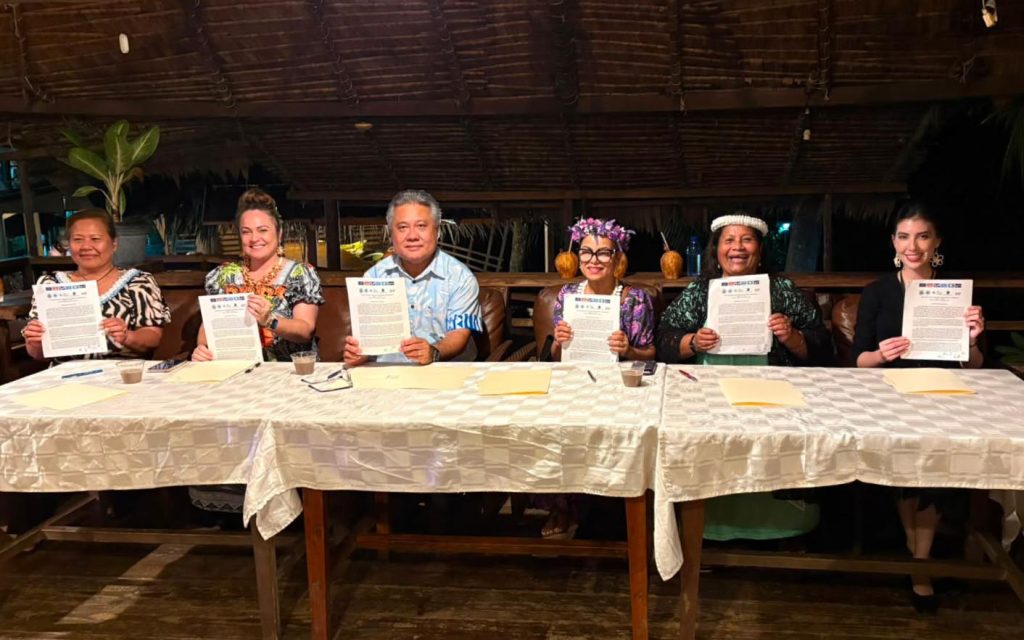 Sarah sits at a table with 5 fellow ministers at the signing of their declaration, holding up their papers.