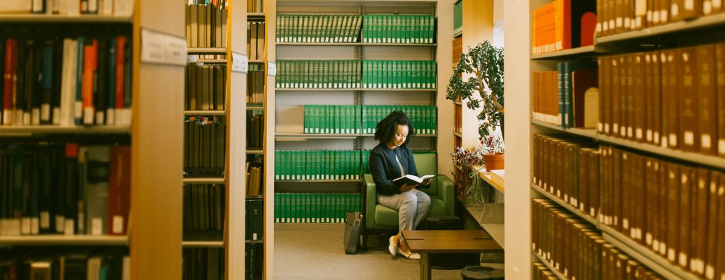Student sitting by a window reading a book in the library surrounded by law books in stacks