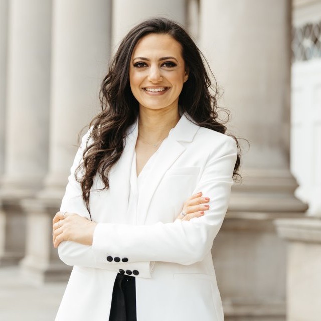 Omnia Shedid stands in front of columns smiling and wearing a white suit jacket.