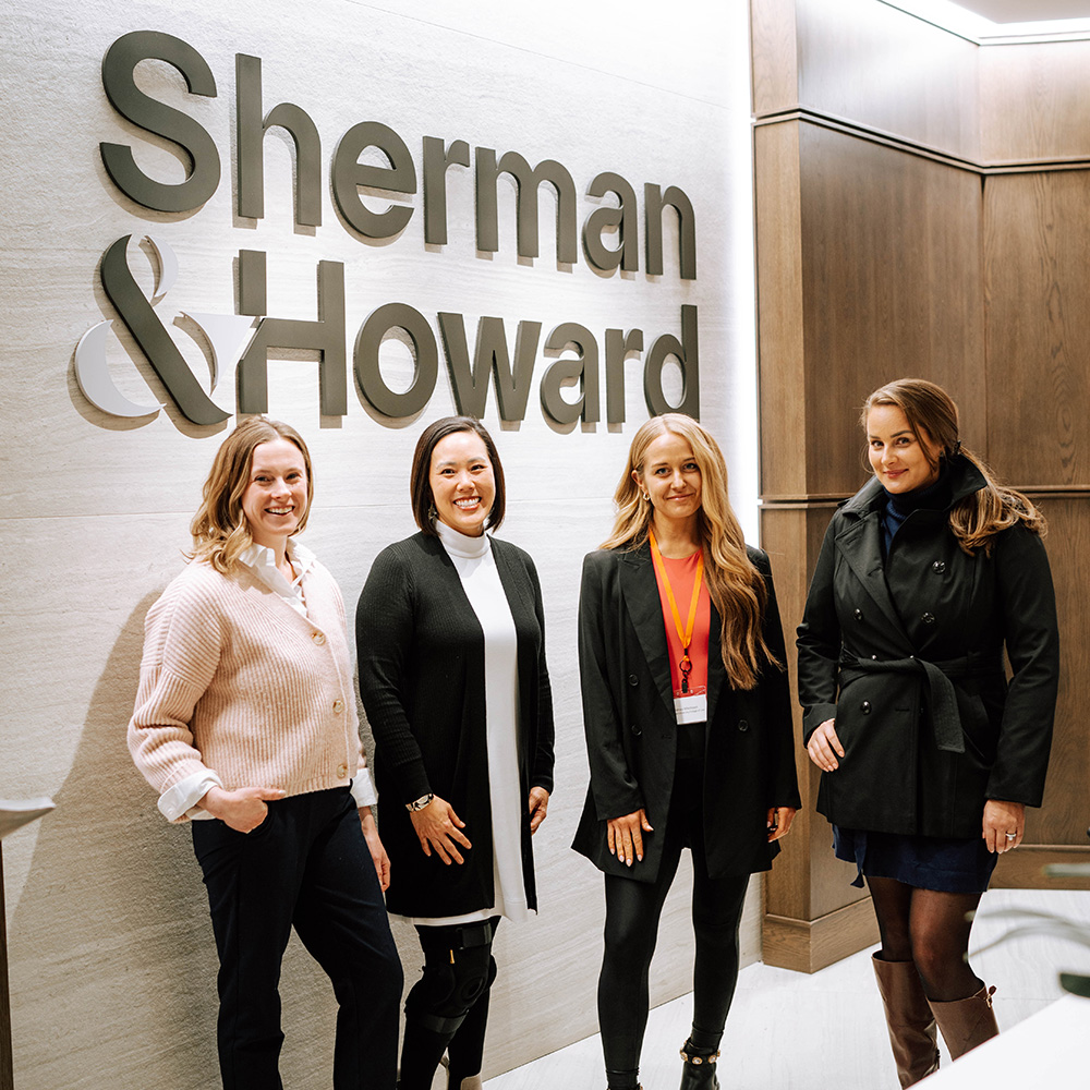 four students stand in front of a Sherman & Howard sign and smile