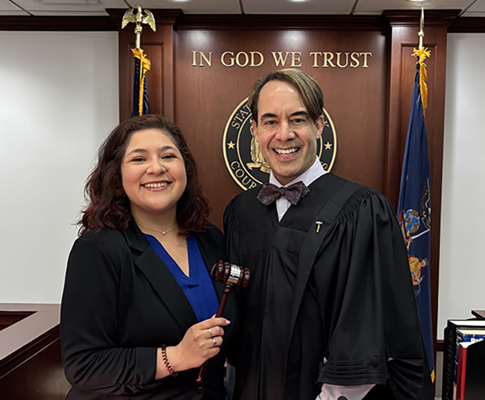 Rebeca Chavar smiles in a courtroom with a judge in front of two flags and an "In God We Trust" sign.