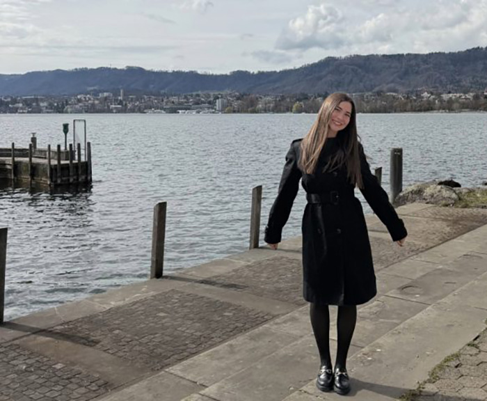 Olivia, a JDi student, stands in front of a body of water in Switzerland wearing a black coat and smiling.
